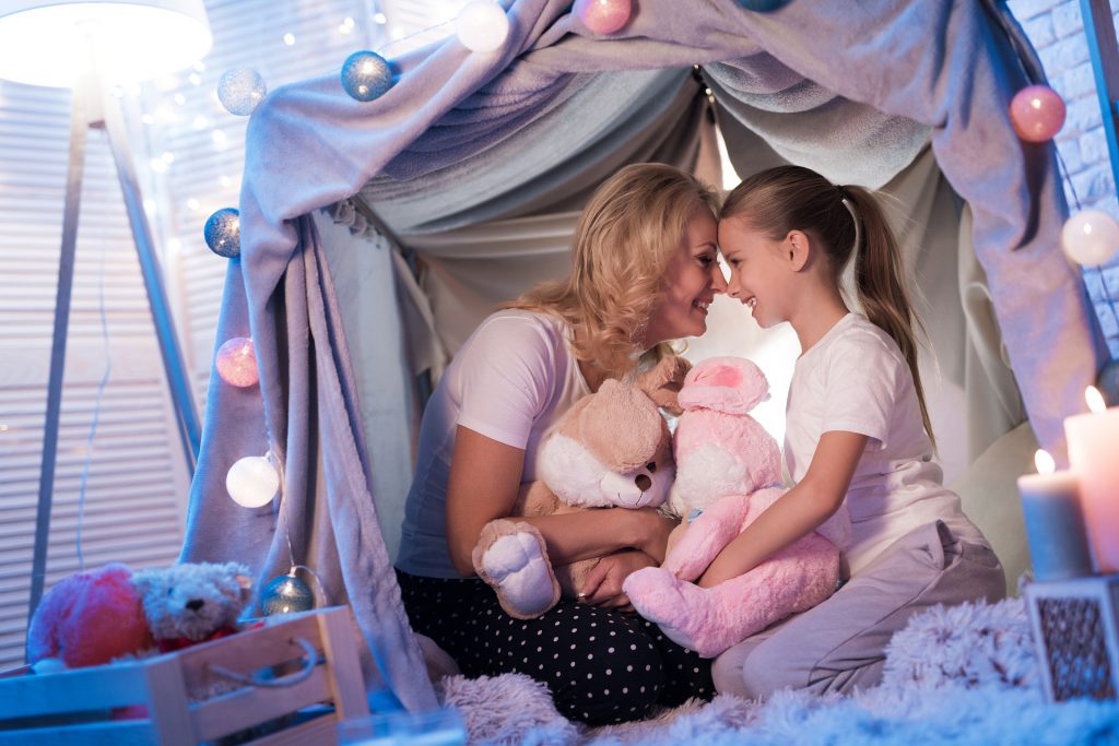 Mom and daughter in a blanket fort with stuffed animals.
