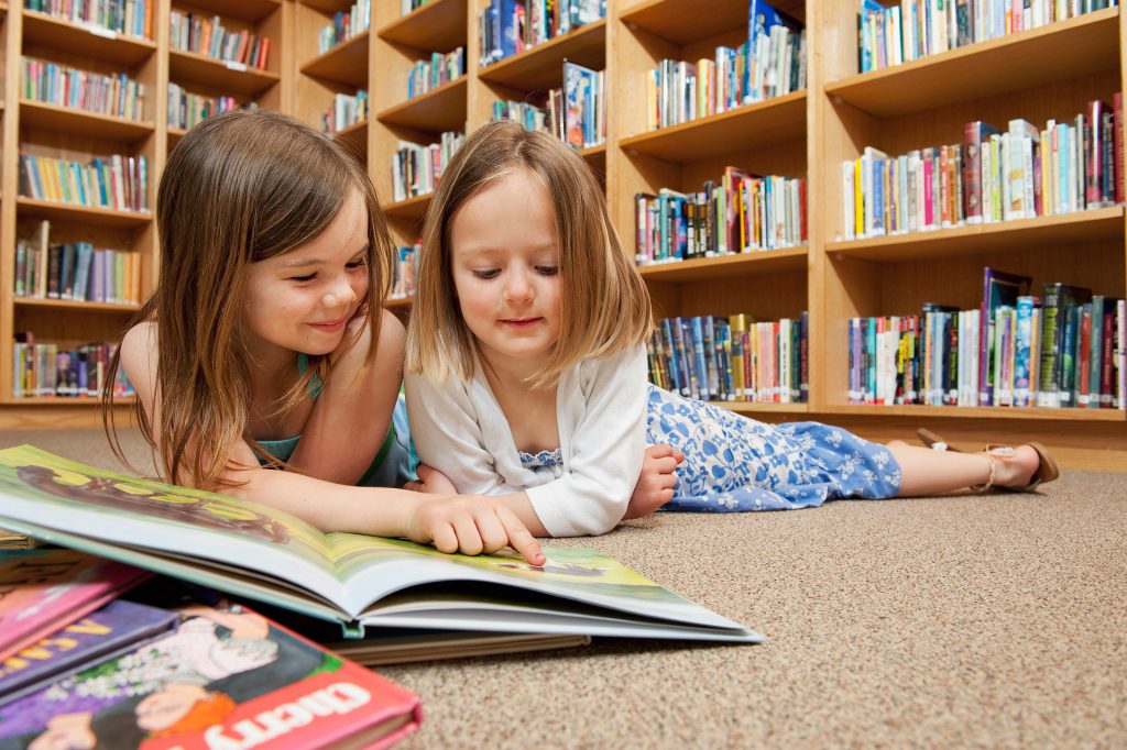 Two girls on the floor of a library with a book. 